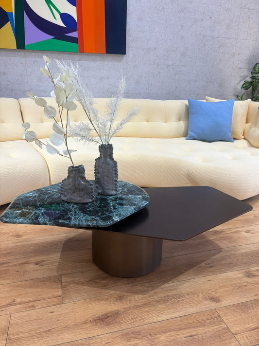 Table basse design contemporaine avec plateau asymétrique noir mat et effet marbre vert foncé, posée sur un pied cylindrique central dans un salon moderne.
