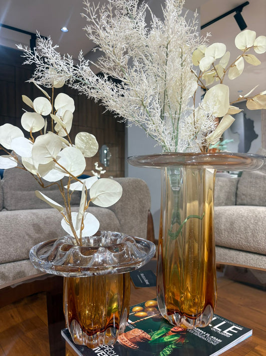 Vases en verre ambré au design sculptural avec ouverture ondulée, décorés de feuillage blanc, posés sur une table dans un salon moderne et élégant.