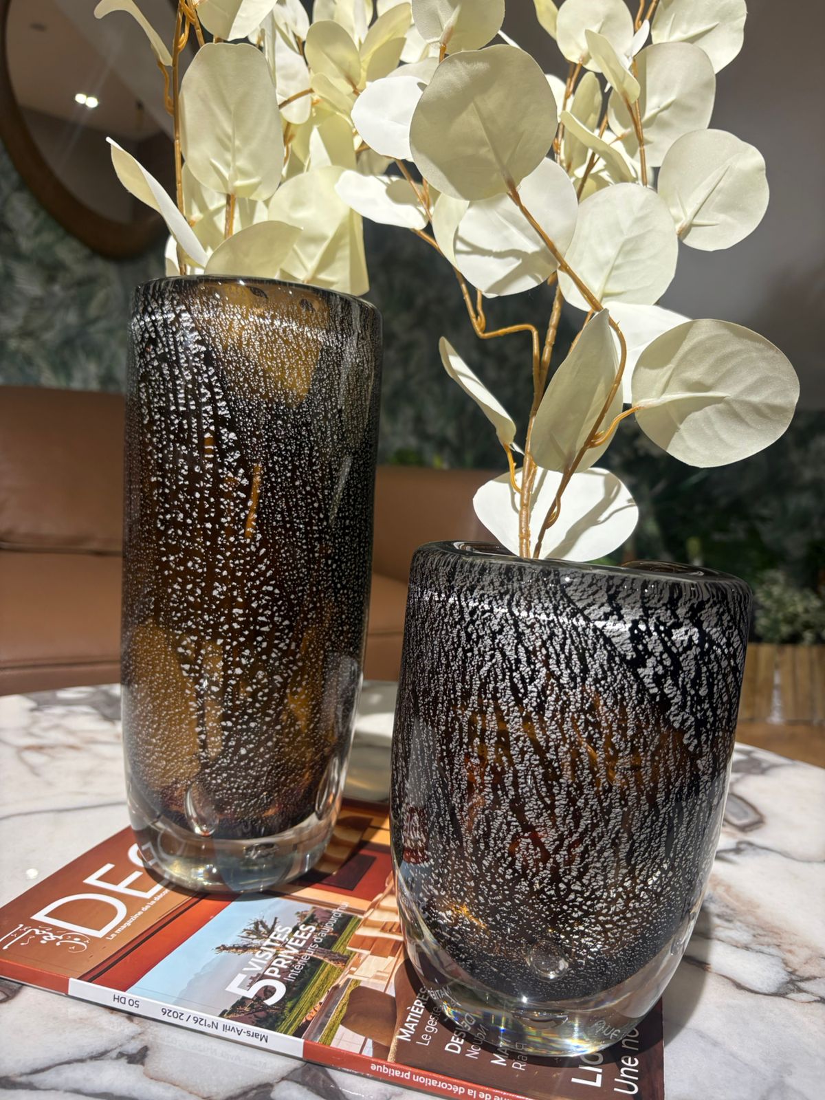 Vases en verre texturé noir et ambré avec effet moucheté, décorés de feuillage blanc, posés sur une table en marbre dans un intérieur moderne et élégant.