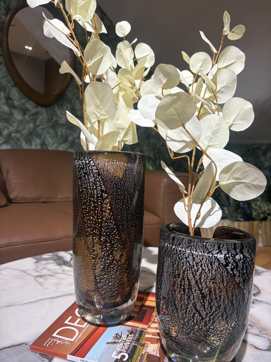 Vases en verre texturé noir et ambré avec effet moucheté, décorés de feuillage blanc, posés sur une table en marbre dans un intérieur moderne et élégant.